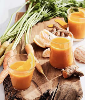 Glasses of Urban Remedy Cold Crusher Juice on a wooden board with carrots, turmeric, and orange slices. 